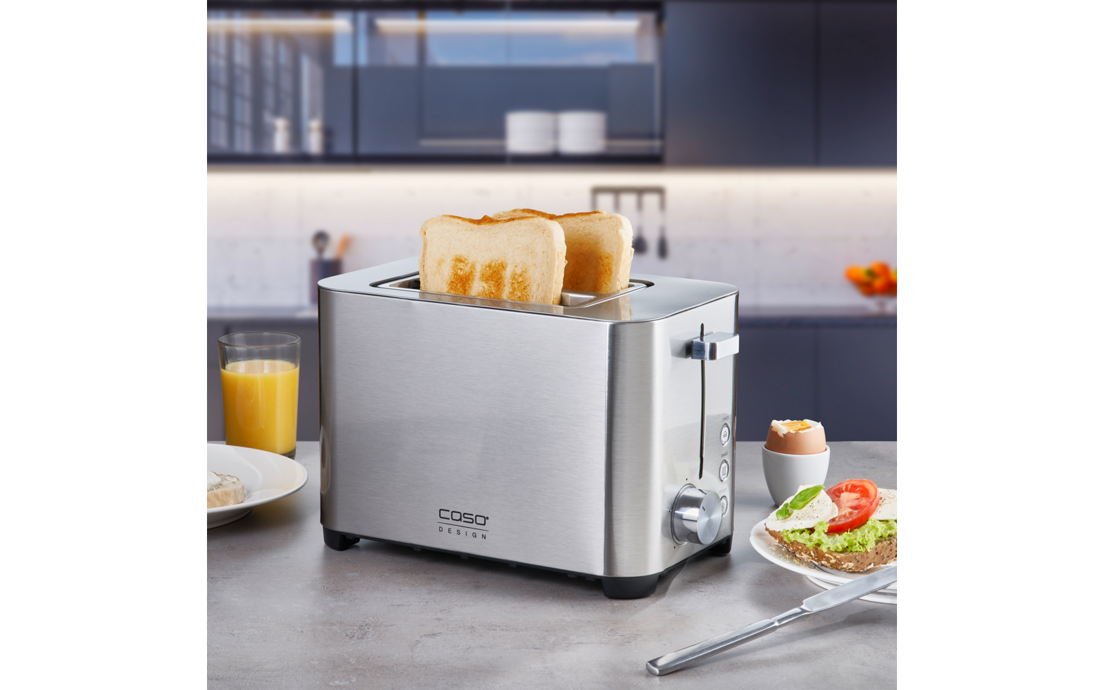 Edelstahl-Toaster mit zwei halb eingesteckten Toastscheiben auf einer Arbeitsplatte; daneben ein Glas Orangensaft, ein Teller mit Brot belegt mit Avocado und einer Tomatenscheibe sowie ein Ei im Eierbecher