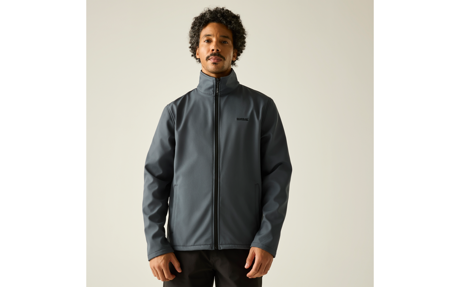 Person mit dunklen, lockigen Haaren trägt eine dunkelgraue Jacke mit durchgehendem Reißverschluss, steht frontal vor hellem Hintergrund