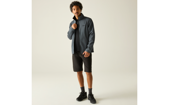 Mann steht vor hellem Hintergrund, trägt graublaue Jacke, schwarzes T‑Shirt, schwarze Shorts, schwarze Socken und schwarze Schuhe; rechte Hand hält den Kragen der Jacke