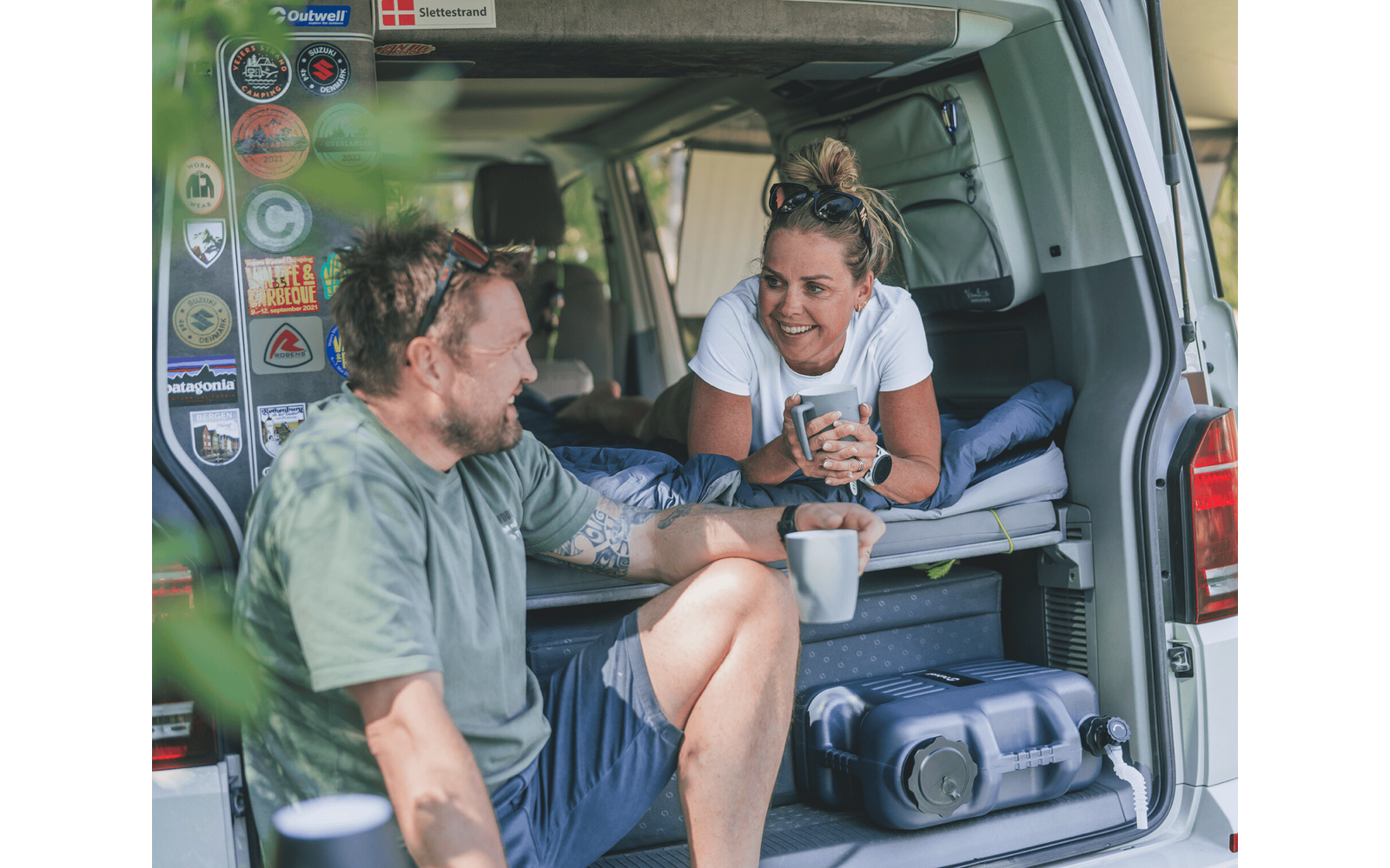 Zwei Personen am geöffneten Heck eines Campingbusses: eine Frau liegt im Innenraum auf einer Schlafplattform, hält eine Tasse und lächelt; ein Mann sitzt am Einstieg und hält ebenfalls eine Tasse. Koffer, Aufkleber an der Tür und verschwommenes Grün 