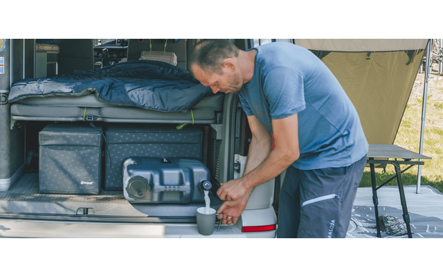Mann am offenen Heck eines Fahrzeugs gießt aus einem Thermobehälter Flüssigkeit in einen Becher; im Heck liegen Schlafsack, Aufbewahrungsboxen und Ausrüstung, daneben ein aufgebautes Zelt