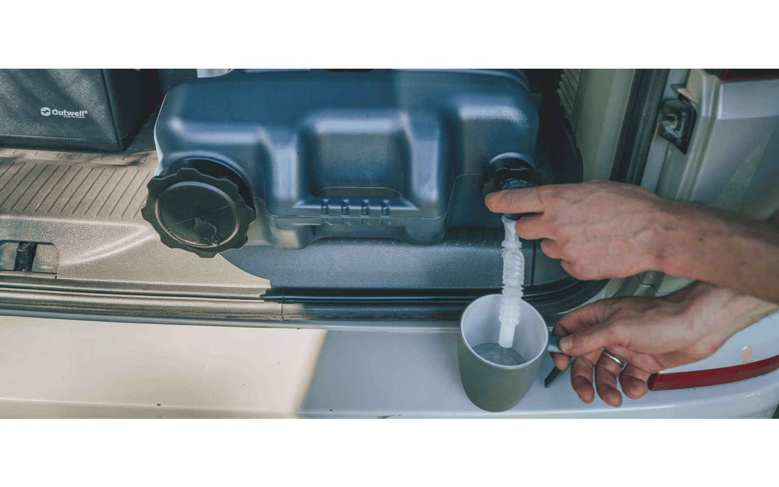 Hand drückt Auslauf eines grauen Kunststoffbehälters im Kofferraum; klare Flüssigkeit fließt in einen Becher