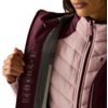 Nahaufnahme des Oberkörpers einer Person, die eine burgundrote Außenjacke aufhält; darunter eine hellrosa gesteppte Innenjacke mit Reißverschluss; weiße Piktogramme auf der Innenseite der Außenjacke; Hand hält die Klappe; Mund und Kinn sowie heller H