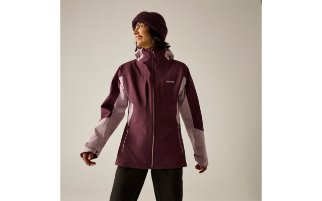 Person in bordeauxfarbener Outdoorjacke mit hellrosa Einsätzen, Strickmütze und schwarzer Hose, schaut zur Seite und lächelt
