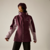 Person in bordeauxfarbener Outdoorjacke mit hellrosa Einsätzen, Strickmütze und schwarzer Hose, schaut zur Seite und lächelt