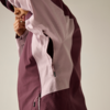 Teilansicht einer Person, die eine zweifarbige Jacke (hellrosa und bordeaux) trägt; ein Arm ist angehoben, Hand am Kinn; seitlicher Reißverschluss und Reißverschlusstasche sichtbar; heller einfarbiger Hintergrund