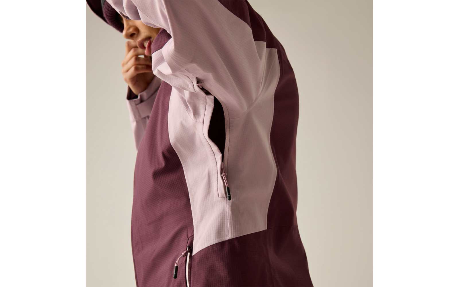 Teilansicht einer Person, die eine zweifarbige Jacke (hellrosa und bordeaux) trägt; ein Arm ist angehoben, Hand am Kinn; seitlicher Reißverschluss und Reißverschlusstasche sichtbar; heller einfarbiger Hintergrund