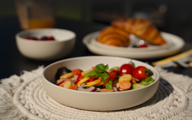 Schale mit Salat (Cherrytomaten, Gurkenscheiben, Möhrenscheiben, rote Zwiebelringe, Basilikumblätter) auf einem geflochtenen Platzset; im Hintergrund unscharf Teller mit Croissants, kleine Schale mit Beeren und Glas mit orangener Flüssigkeit auf dunk