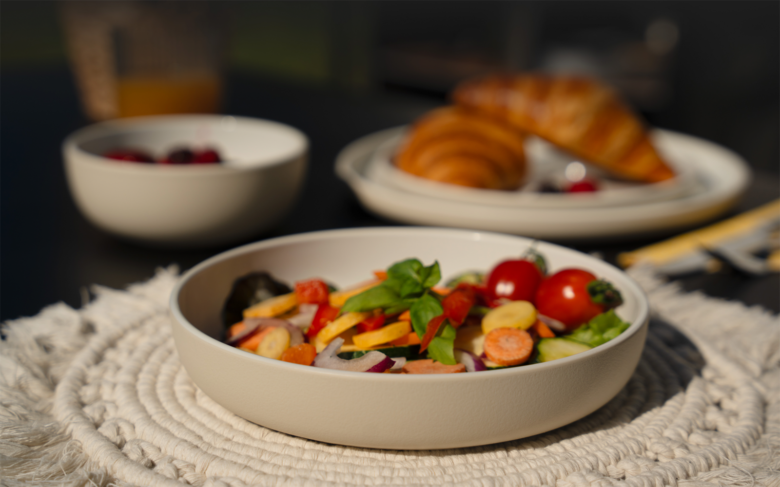 Schale mit Salat (Cherrytomaten, Gurkenscheiben, Möhrenscheiben, rote Zwiebelringe, Basilikumblätter) auf einem geflochtenen Platzset; im Hintergrund unscharf Teller mit Croissants, kleine Schale mit Beeren und Glas mit orangener Flüssigkeit auf dunk