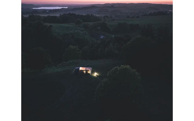 Dämmerungslandschaft mit dunklen Bäumen und einer kleinen Straße, auf der ein beleuchtetes Fahrzeug fährt