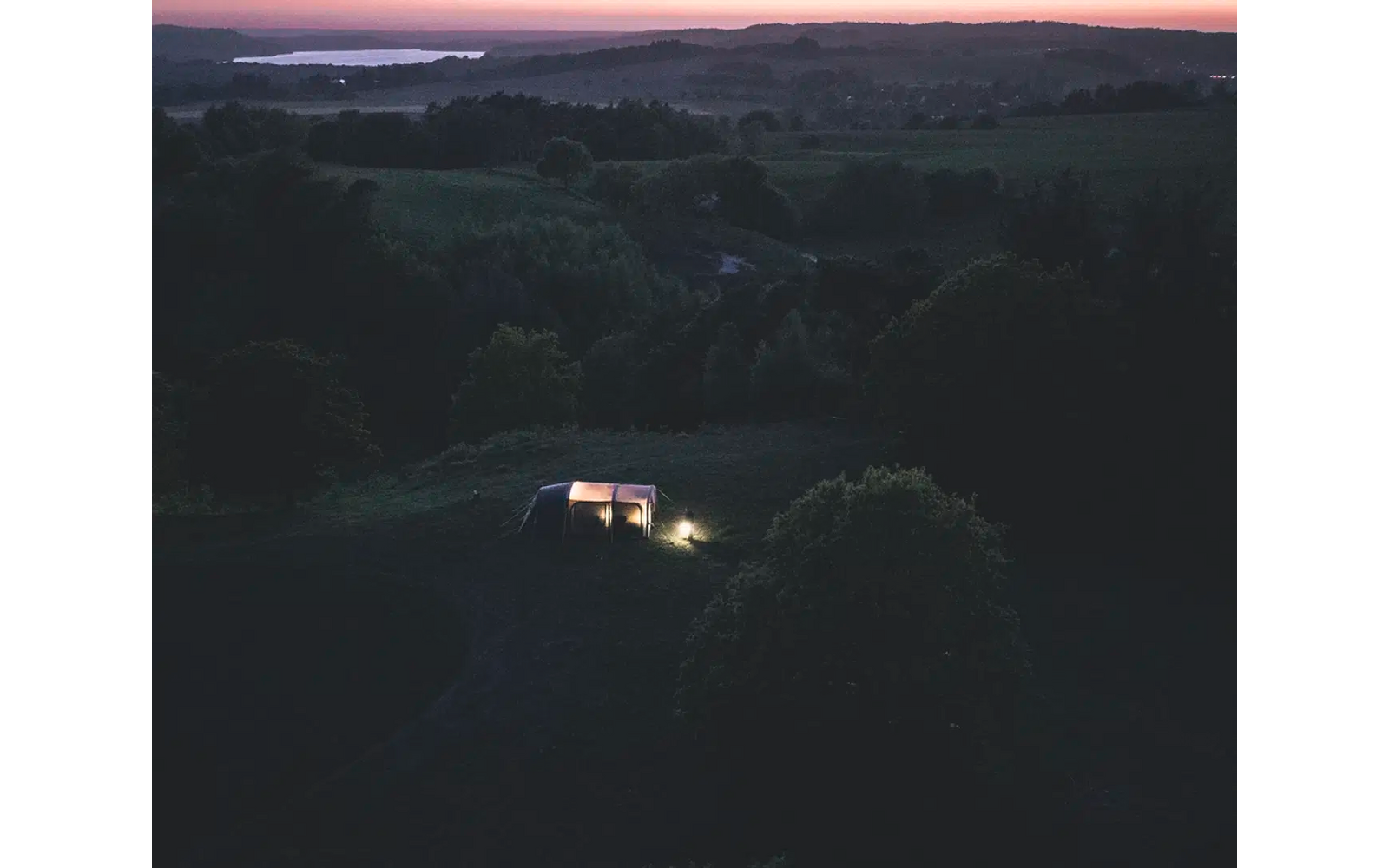 Dämmerungslandschaft mit dunklen Bäumen und einer kleinen Straße, auf der ein beleuchtetes Fahrzeug fährt