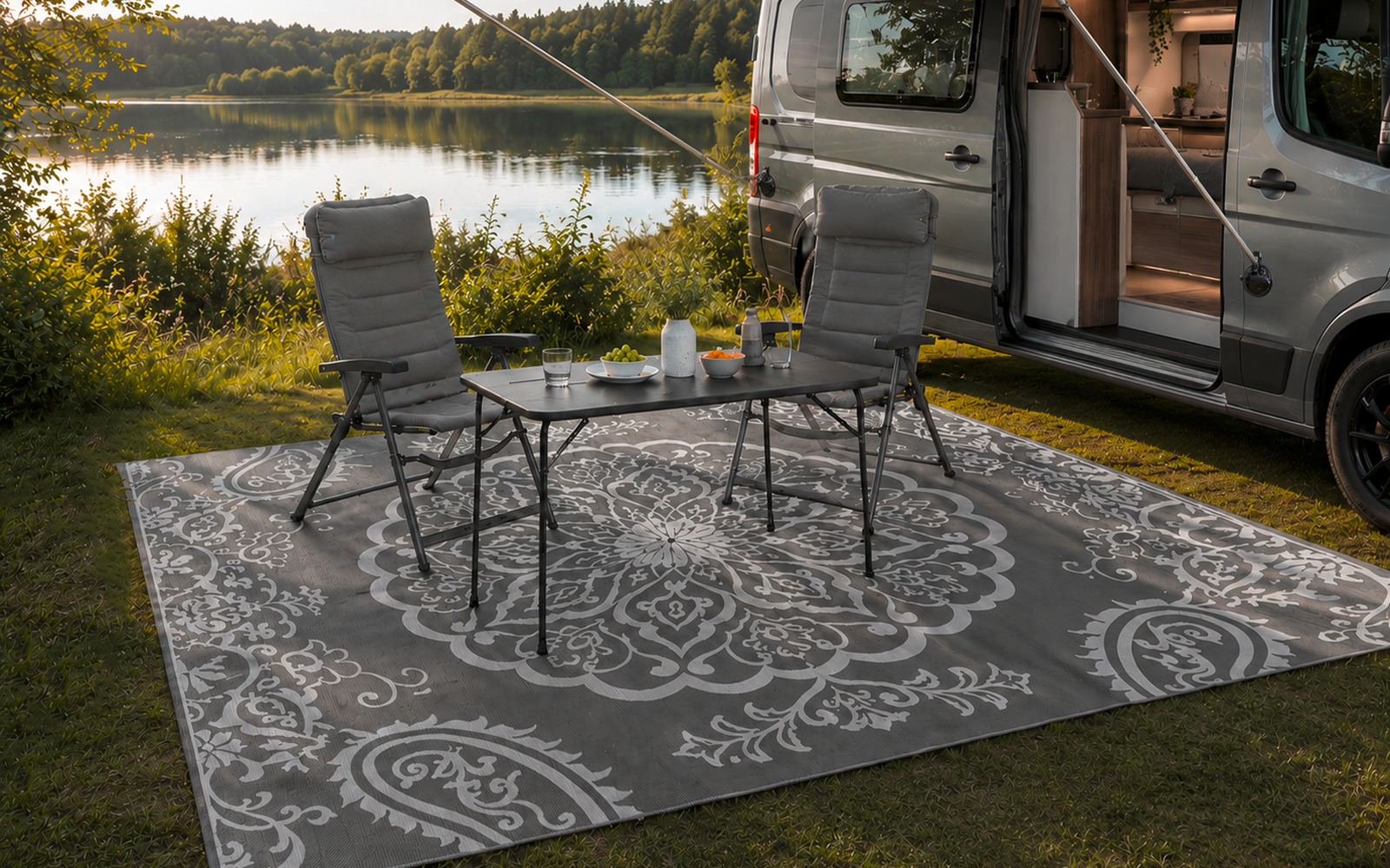 Fourgon aménagé gris avec porte latérale ouverte et auvent déployé ; devant un tapis d'extérieur à motif, deux chaises de camping pliantes et une petite table avec une carafe et des verres, au bord d'un lac avec des arbres en arrière-plan
