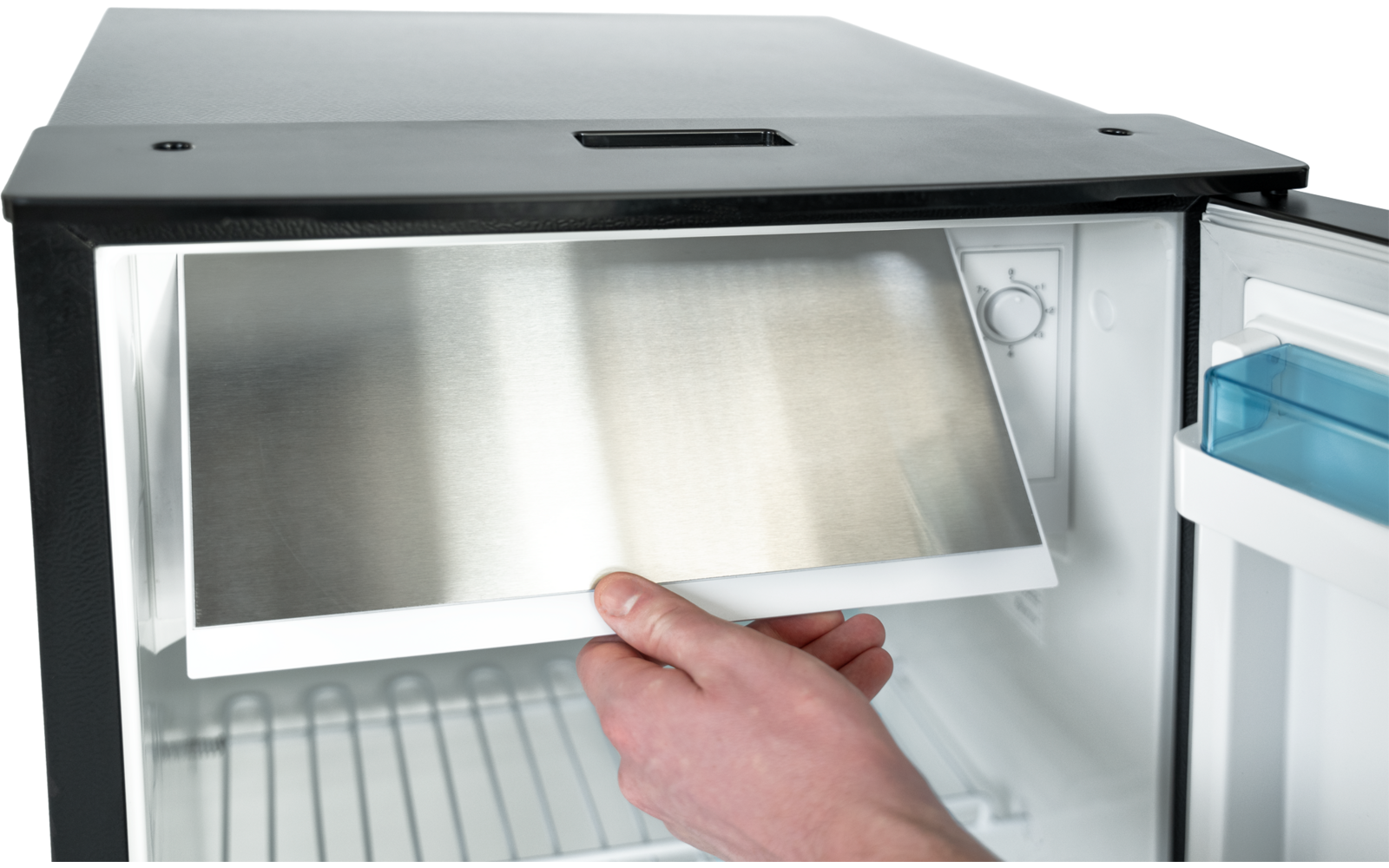 Hand hält ein rechteckiges Edelstahlblech im Inneren eines geöffneten kleinen Kühlschranks; Drahtgitterboden und blaue Türablage sichtbar