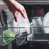 Une main tient un verre transparent au-dessus du panier ouvert du lave-vaisselle; plusieurs verres transparents, un gobelet en plastique vert et des assiettes blanches dans le panier