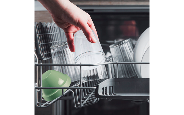 Une main tient un verre transparent au-dessus du panier ouvert du lave-vaisselle; plusieurs verres transparents, un gobelet en plastique vert et des assiettes blanches dans le panier