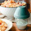 Récipient plastique transparent avec couvercle turquoise contenant un liquide blanc ; au premier plan une assiette de macaroni ; en arrière-plan un bol blanc de macaroni avec petits morceaux orange et rouges sur une table en bois