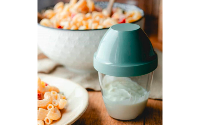 Récipient plastique transparent avec couvercle turquoise contenant un liquide blanc ; au premier plan une assiette de macaroni ; en arrière-plan un bol blanc de macaroni avec petits morceaux orange et rouges sur une table en bois