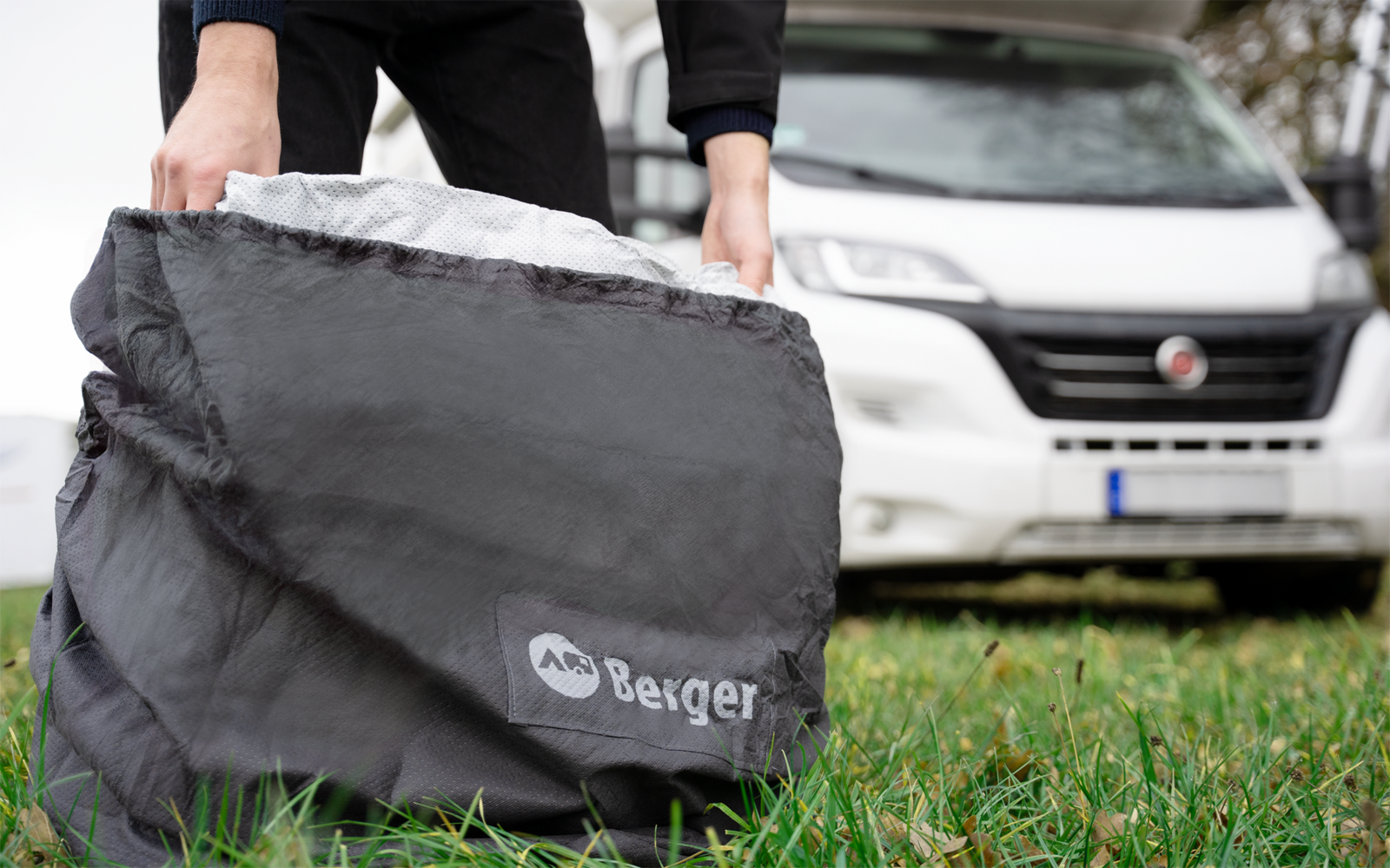 Hands hold a black bag with a white 'Berger' logo on grass in front of a white van