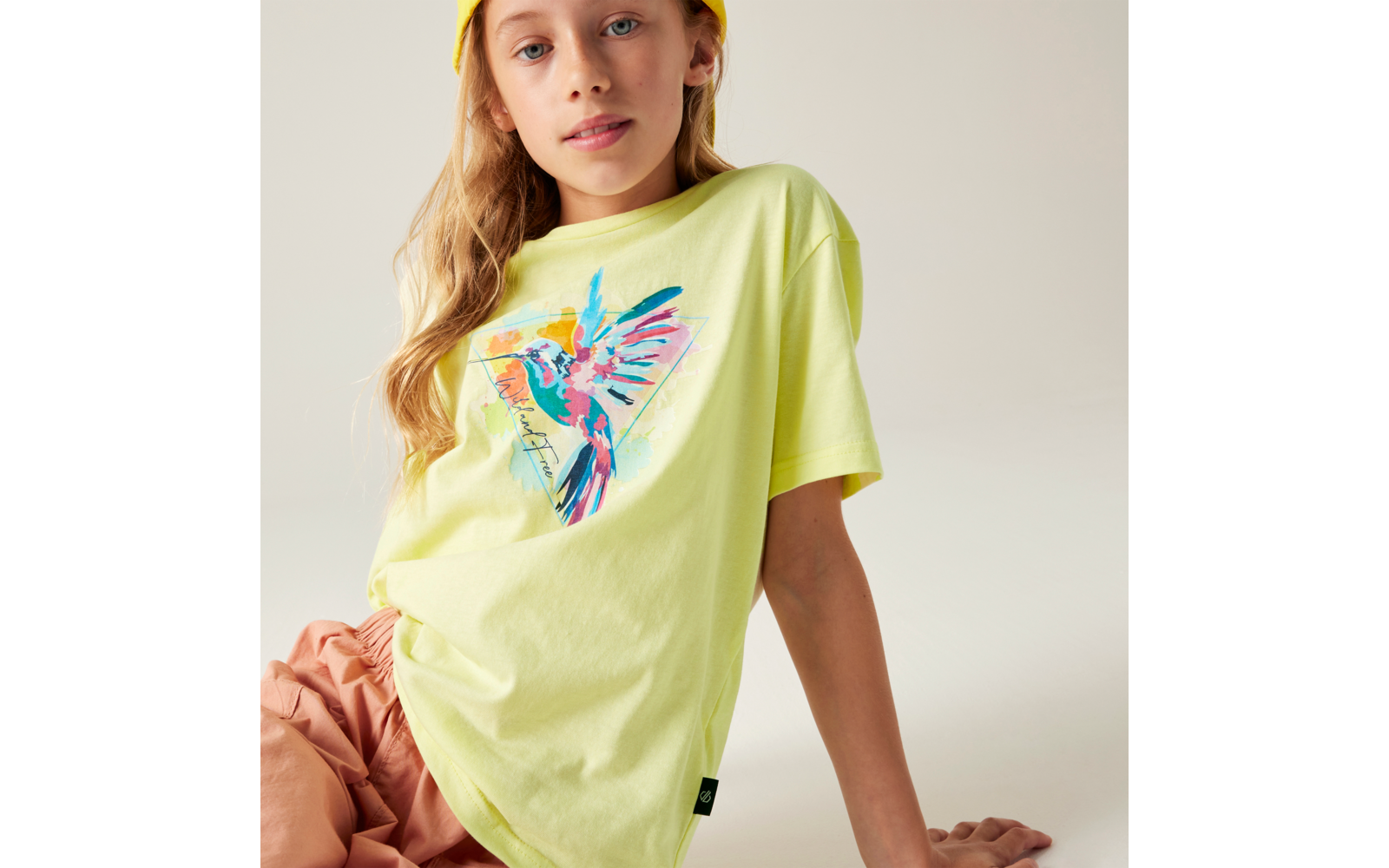 Person sitzt leicht seitlich, trägt hellgelbes T‑Shirt mit buntem Kolibri‑Motiv, gelbe Mütze und rosafarbene Hose vor hellem Hintergrund