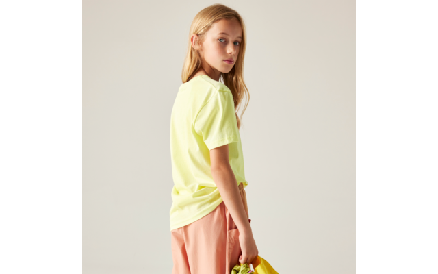 Seitliche Ansicht einer Person mit blonden Haaren, gelbem T‑Shirt und pfirsichfarbenen Shorts, die eine gelbe Tasche hält vor hellem Hintergrund