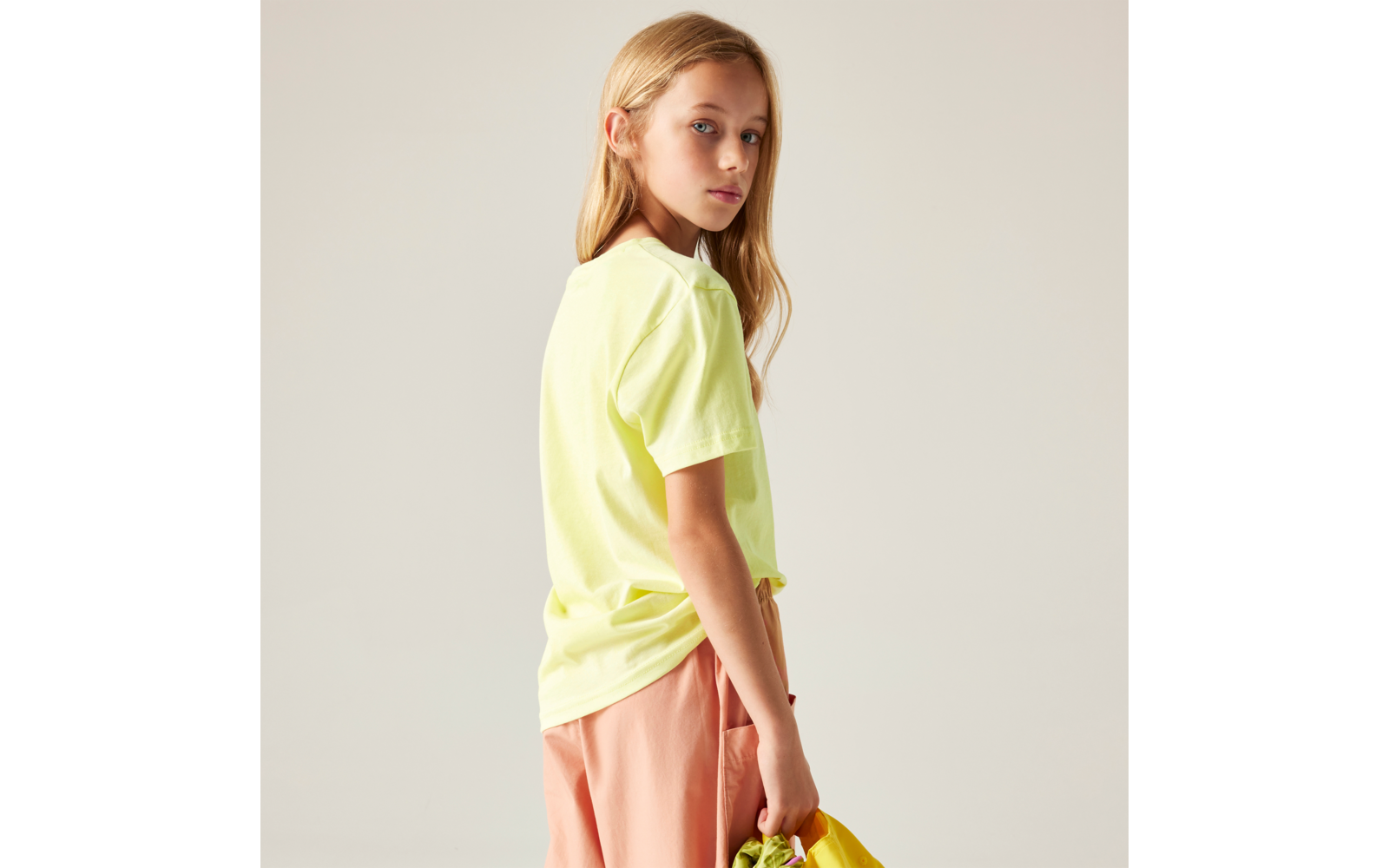 Seitliche Ansicht einer Person mit blonden Haaren, gelbem T‑Shirt und pfirsichfarbenen Shorts, die eine gelbe Tasche hält vor hellem Hintergrund