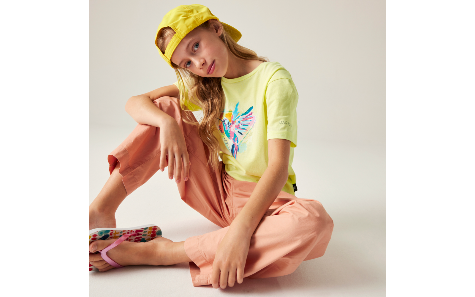 Sitzendes Mädchen auf hellem Hintergrund, trägt gelbe Schirmmütze, gelbes T‑Shirt mit buntem Vogelmotiv, lachsfarbene Hose und bunte Sandalen