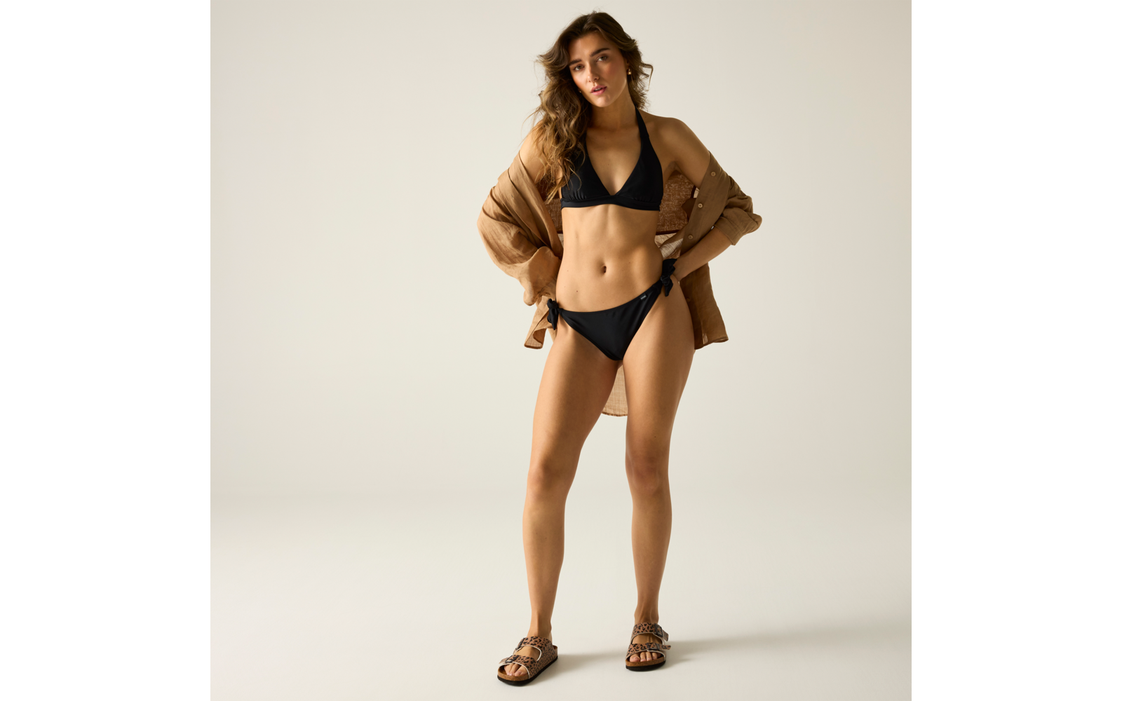 Person in schwarzem Bikini und offenem braunen Jackett, steht vor hellem Hintergrund, trägt braune Sandalen