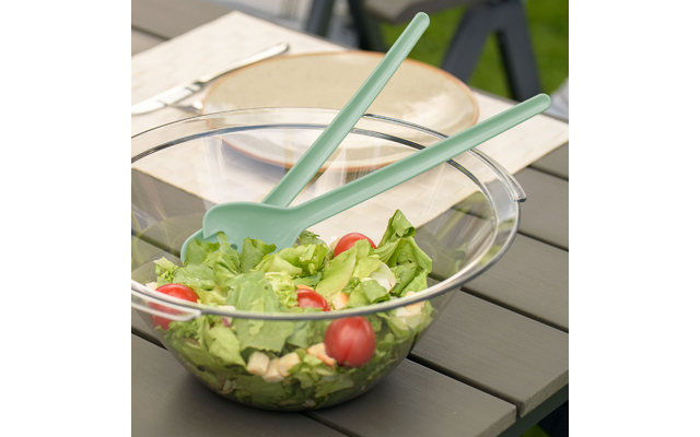 Saladier en verre transparent avec feuilles de salade, tomates cerises, petits dés et deux couverts à salade vert menthe sur une table de jardin grise ; assiettes et couverts en arrière-plan