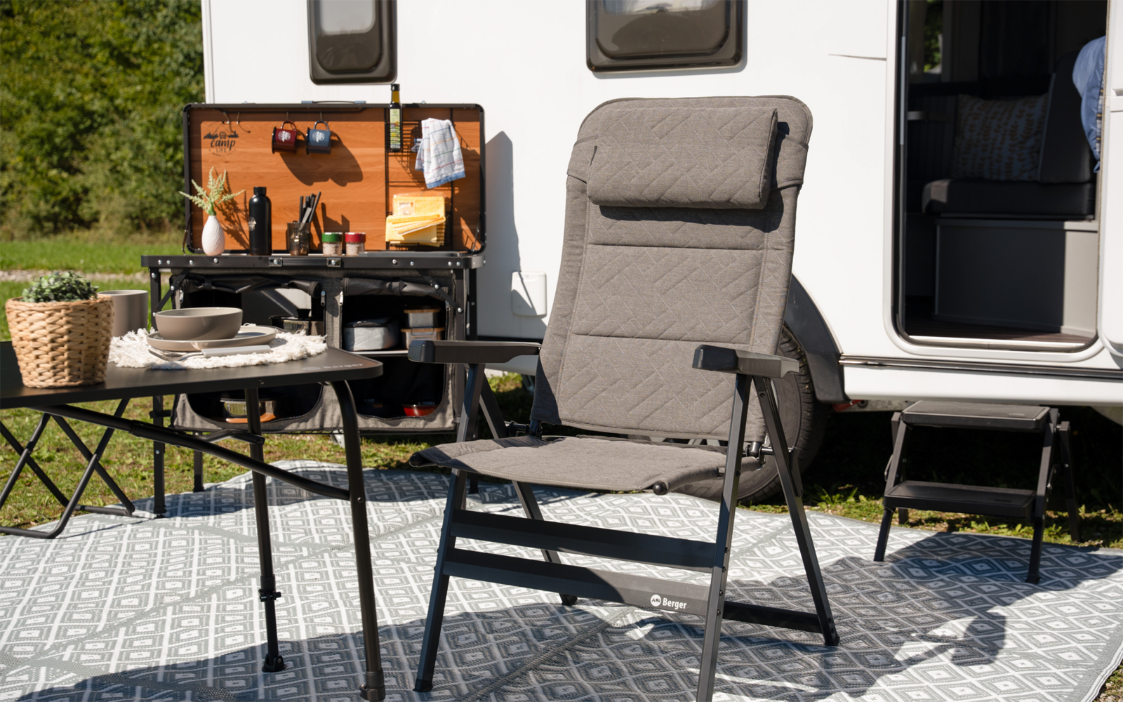 Gepolsterter Campingstuhl vor einem Wohnmobil, kleiner Tisch mit Tellern und Tassen, geöffnete Küchenklappe des Wohnmobils, gemusteter Outdoor-Teppich und Korb auf dem Gras