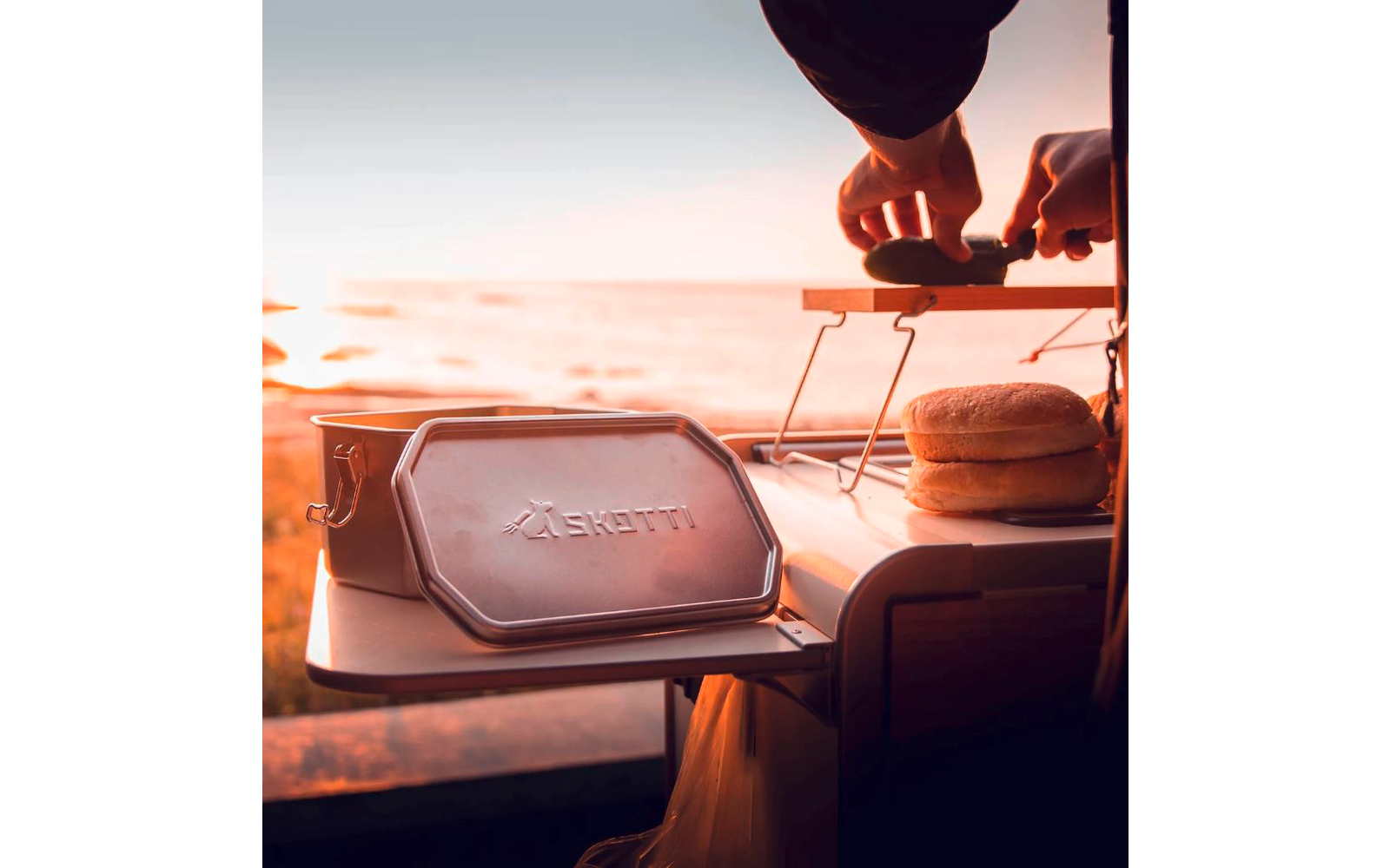Main tourne un petit grill pliant estampillé 'Skotti', un petit pain à côté, en arrière‑plan mer et soleil à l'horizon