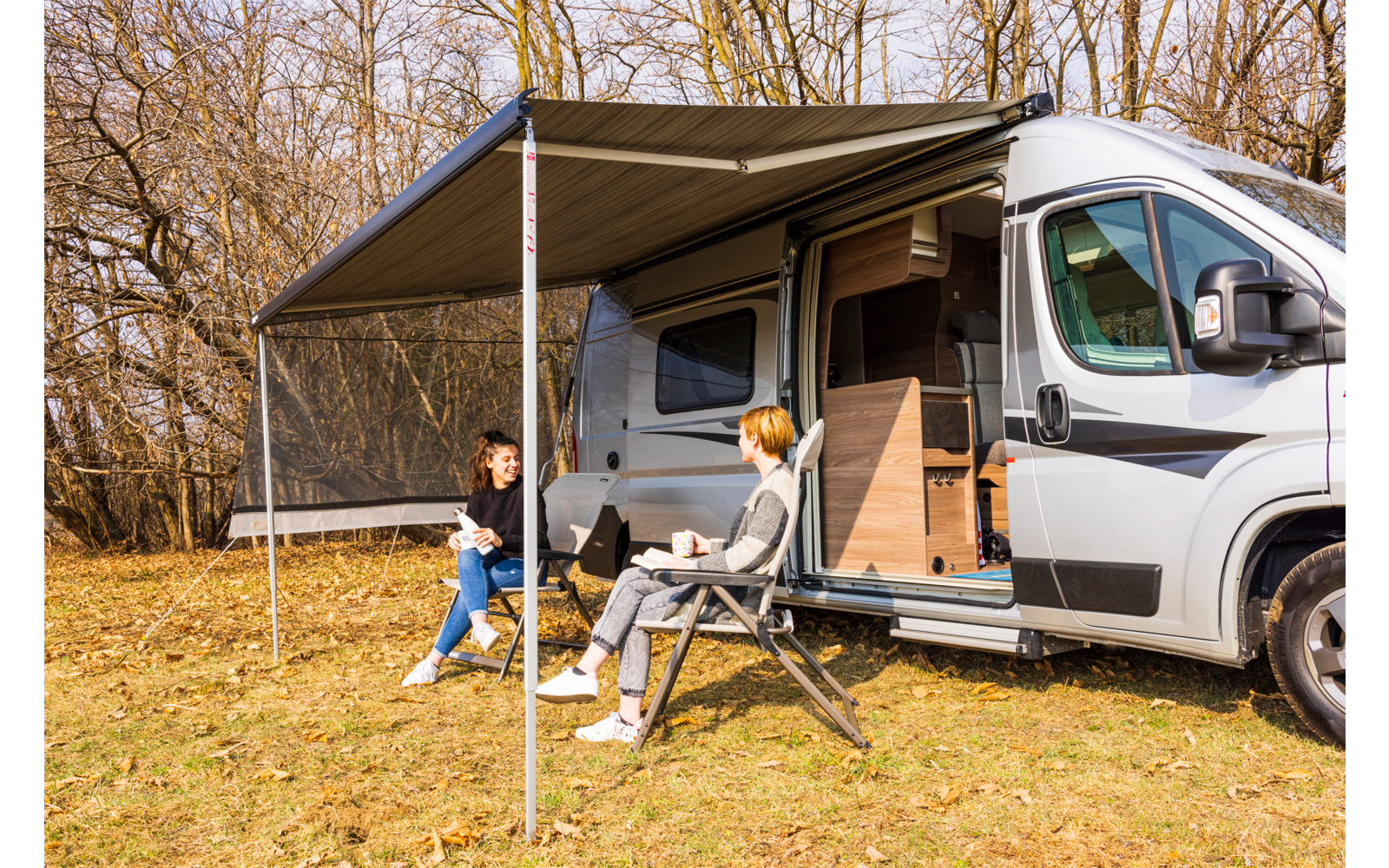 Wohnmobil mit ausgefahrener Markise; zwei Personen sitzen auf Campingstühlen vor dem offenen Seiteneingang; Wiese mit kahlen Bäumen im Hintergrund