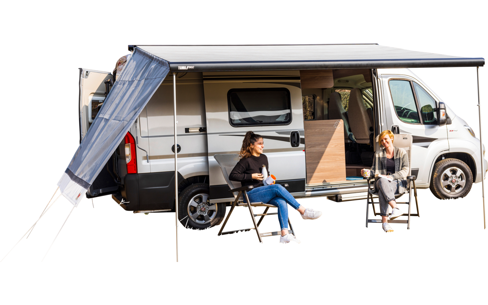 Weißer Camper-Van mit ausgefahrener Markise auf einer Wiese vor kahlen Bäumen; zwei Personen sitzen an einem kleinen Tisch vor der offenen Seitentür