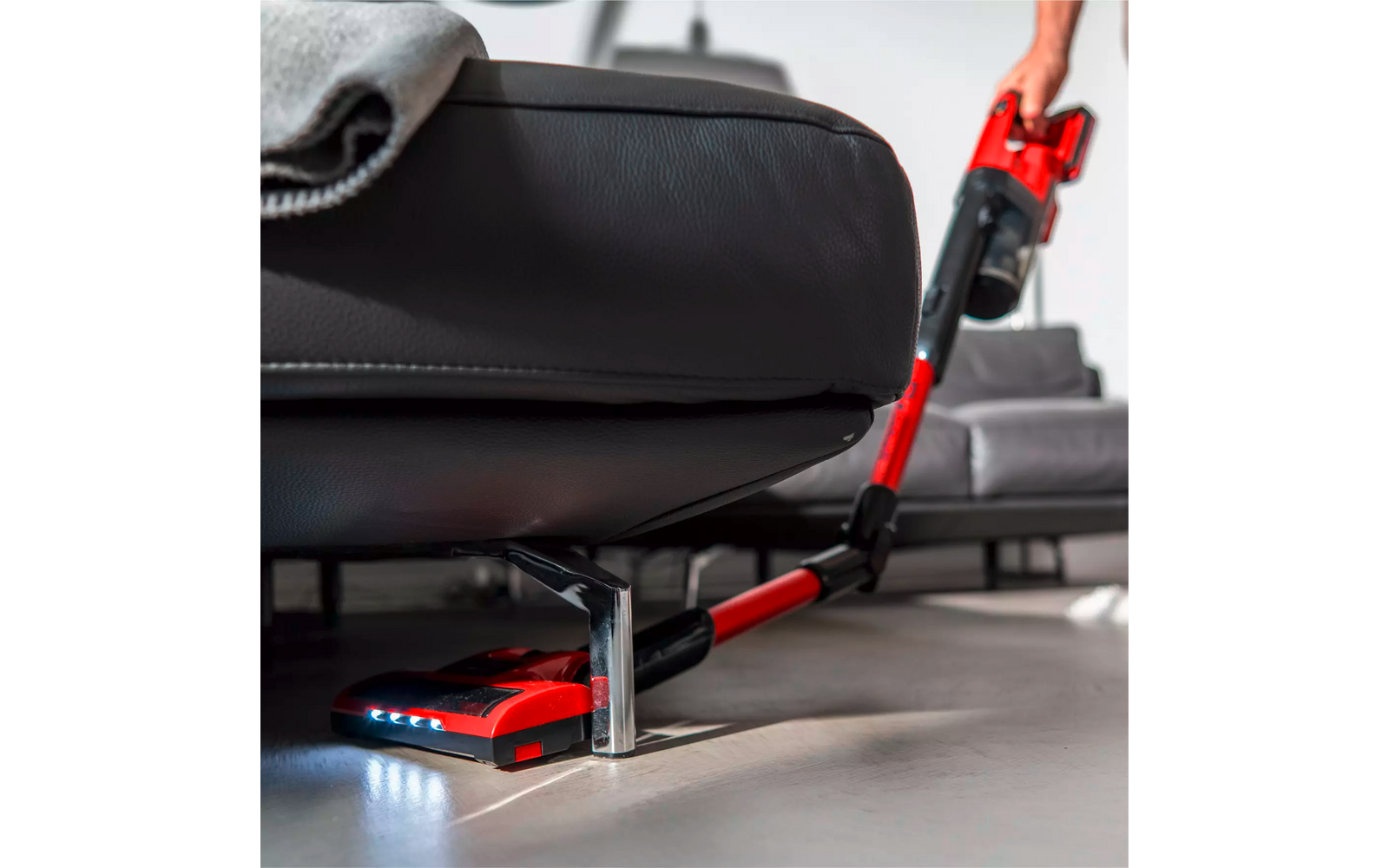 Aspirateur-balai rouge avec éclairage glissé sous un canapé gris en cuir à pieds métalliques; main sur la poignée visible