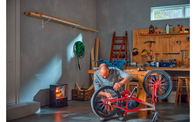 Man kneeling in a workshop repairing an upside-down red bicycle; workbench with tools, ladder, wood stove with visible flame, and a green hose on the wall
