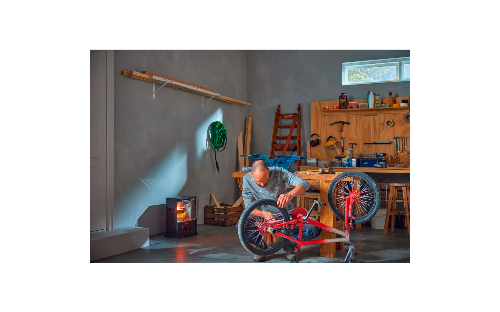 Man kneeling in a workshop repairing an upside-down red bicycle; workbench with tools, ladder, wood stove with visible flame, and a green hose on the wall