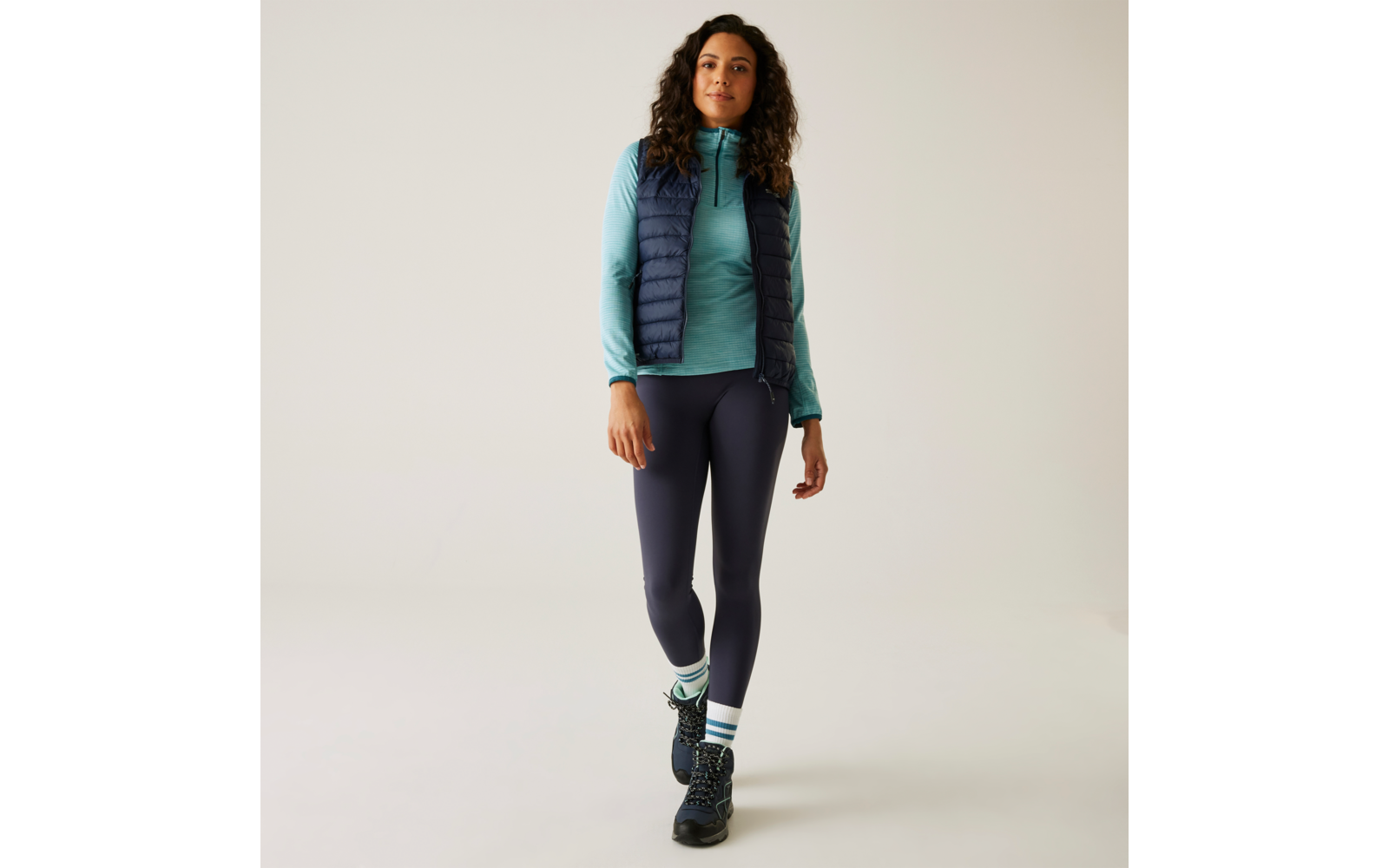 Vrouw met lang krullend haar draagt een doorgestikt vest, turquoise trui, donkere legging en zwarte schoenen, staand en vooruit kijkend