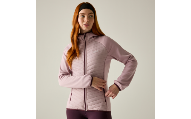 Person with long red hair and black headband wearing a light pink quilted zip-up jacket and dark leggings, one hand at hip, plain light background