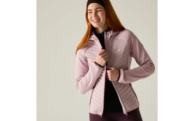 Smiling woman with red hair and headband closing a light pink quilted jacket over a black top against a light background