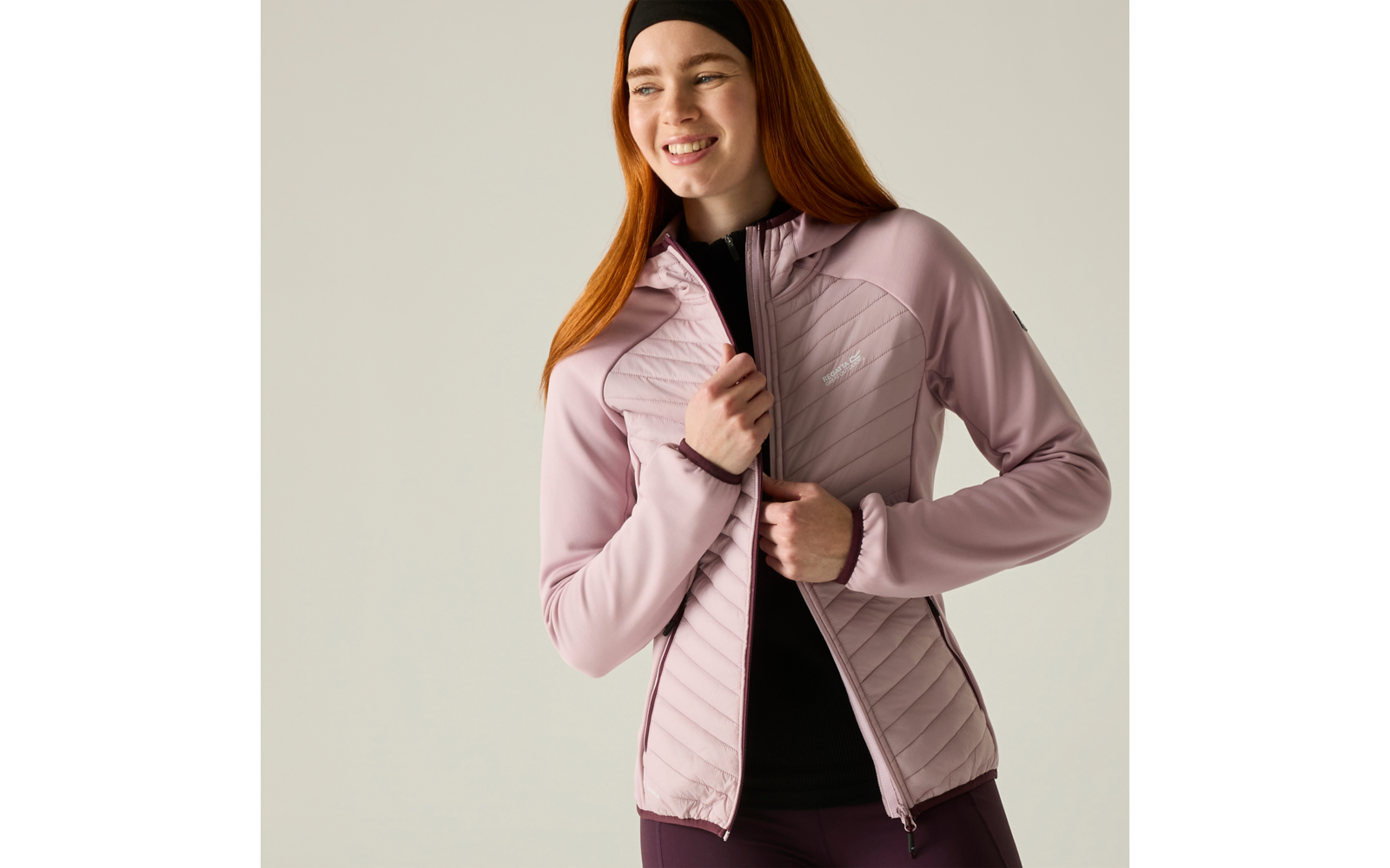 Smiling woman with red hair and headband closing a light pink quilted jacket over a black top against a light background