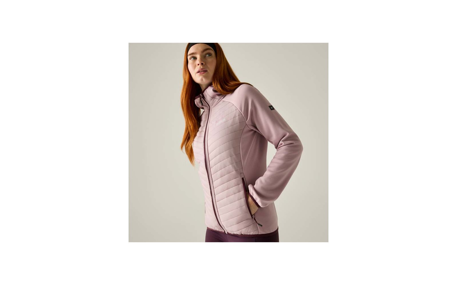 Person with long red hair and a black headband, wearing a light pink quilted zip jacket, hands in side pockets, light background
