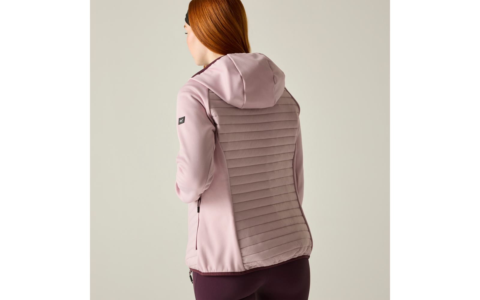 Person seen from back wearing a light pink hooded jacket with a horizontal quilted back panel and dark purple leggings; reddish hair visible