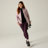 Person with red hair wearing a light pink quilted jacket, black top, dark purple leggings and black sneakers, holding a black accessory in the right hand, light plain background
