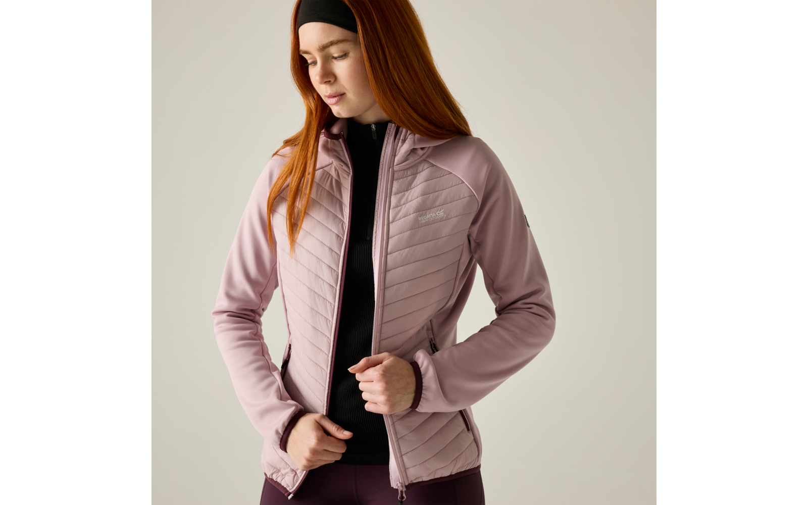 Person with long red hair and black headband fastening a light pink quilted jacket over a black zip-up top