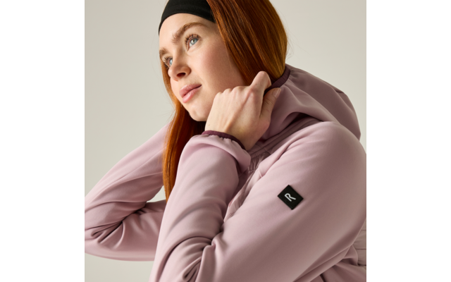 Person with red hair and headband wearing a pink hoodie, adjusting the hood, looking up-right against a light plain background