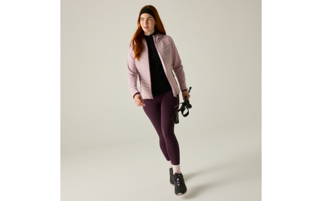 Person with red hair wearing a light pink quilted jacket, black top, dark purple leggings and black sneakers, holding a black accessory in the right hand, light plain background