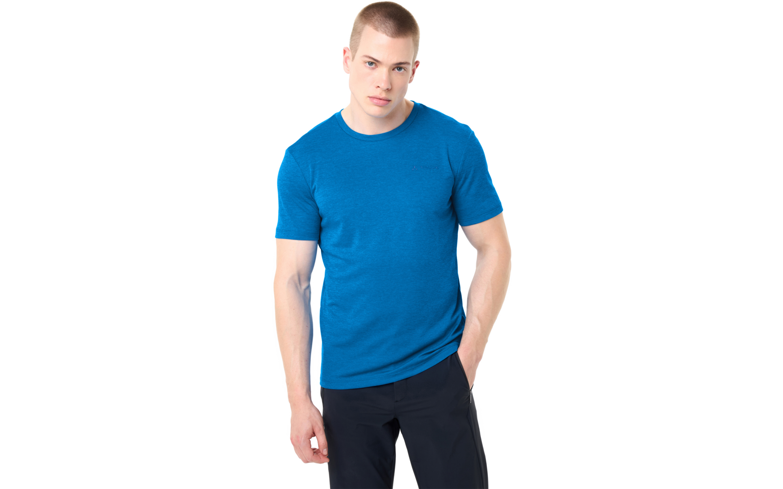 Jonge man met kort haar, draagt een blauw T‑shirt met korte mouwen en een donkere broek, staat voor een witte achtergrond, één hand in de zak