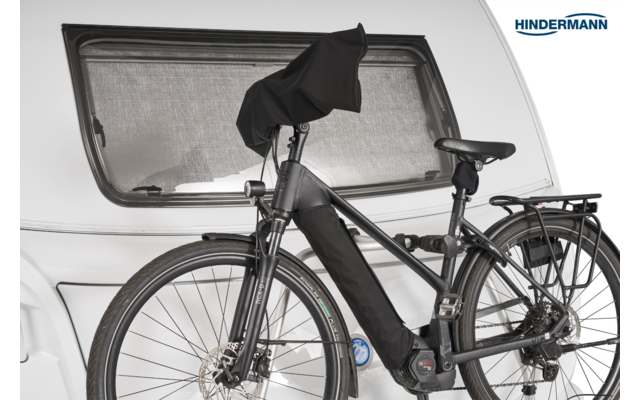Schwarzes Elektrofahrrad von vorn-rechts, Vorder- und Hinterrad sichtbar, Akku am Unterrohr mit schwarzer Hülle, Sattel und Gepäckträger, Lenker teils mit schwarzem Tuch bedeckt, weiße Wohnwagenfront mit Fenster und 'Hindermann'-Logo im Hintergrund