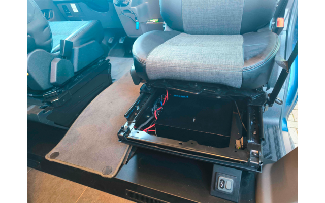 Siège de véhicule dont la partie inférieure est visible ; sous le siège un boîtier noir avec des câbles rouges et noirs fixé sur des rails métalliques, un tapis de sol gris à côté, un autre siège et le sol du véhicule en arrière-plan