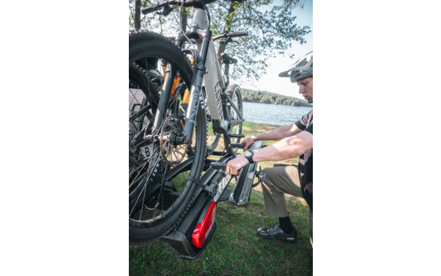 Person mit Fahrradhelm befestigt ein E‑Bike an einem Heckfahrradträger neben einem See