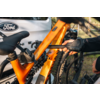 Orangefarbener Mountainbike-Rahmen mit Hinterradfederung, Sattel und grobstolligem Hinterreifen, an einer schwarzen Trägerbefestigung geklemmt; behandschuhte Hand; weißes Auto mit Ford-Logo im Hintergrund